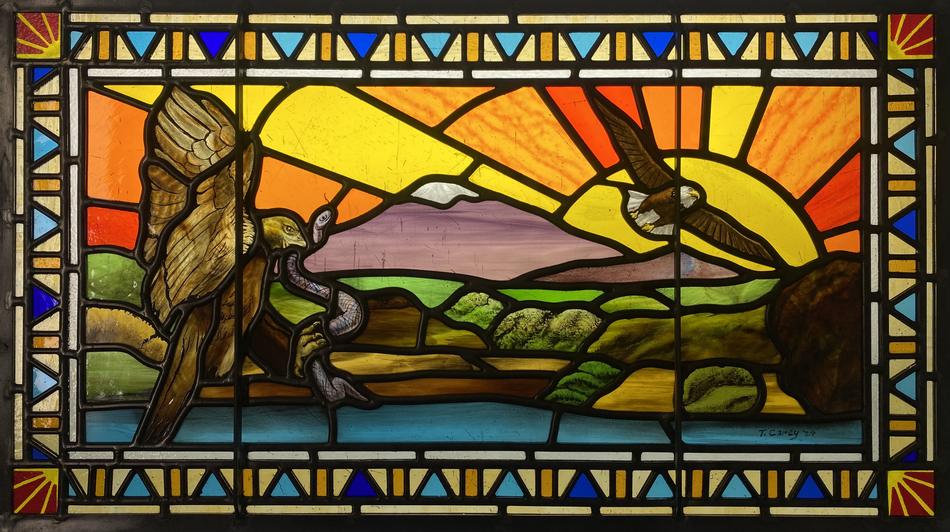 Painted stained glass window depicting two eagles and a mountain landscape at sunset with geometric border